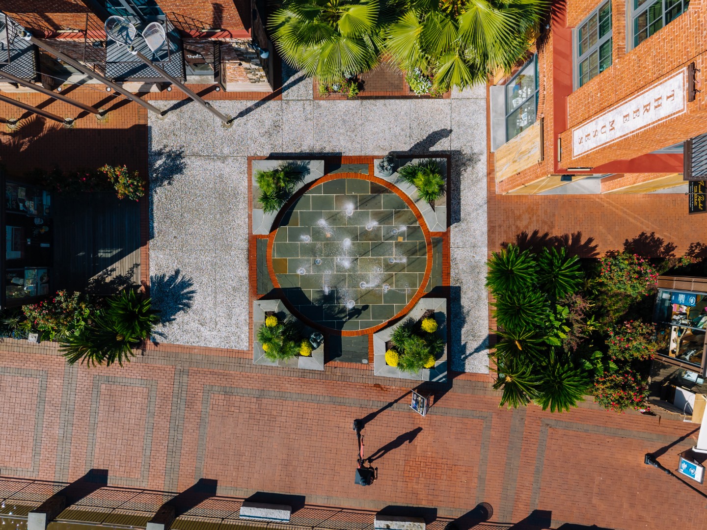 Aerial view of a circular fountain with water jets surrounded by palm trees and benches in a brick-paved plaza, with a person walking nearby and red-roofed buildings on either side.