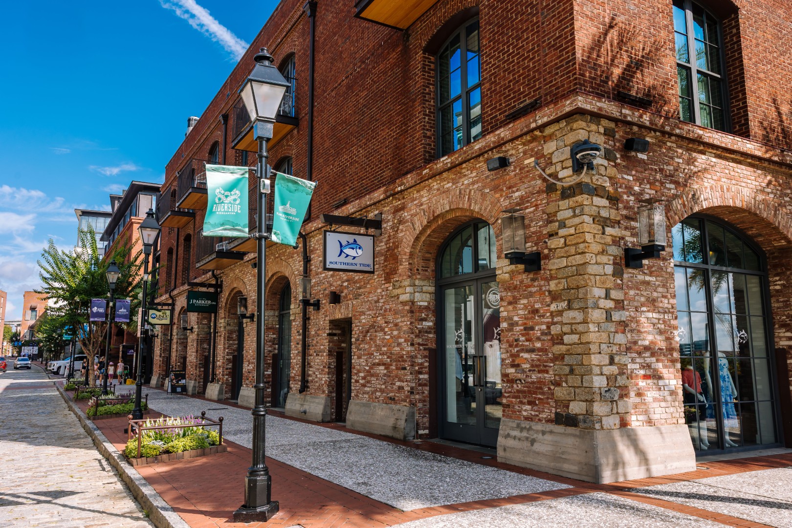 A red brick building with arched windows and storefronts lines a cobblestone street on a sunny day, with green banners hanging from a black lamppost and clear blue sky above.