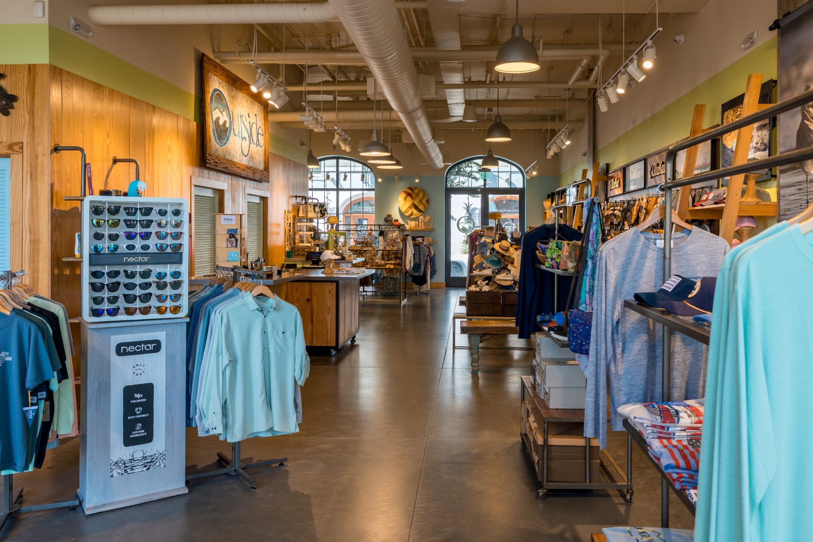 A neatly organized retail store with racks of clothing, sunglasses display, wooden shelves, and various merchandise. Warm lighting and large windows create a welcoming atmosphere.
