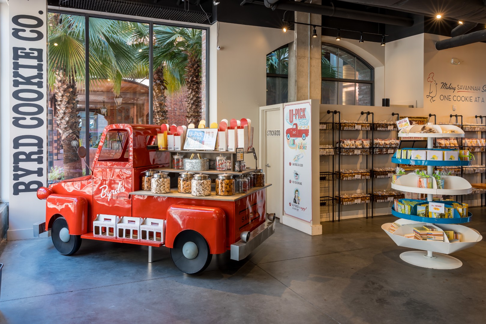 A bright cookie shop features a red truck display loaded with glass jars of cookies, a rotating rack with cookie boxes, and large windows showing palm trees outside. Shelves of packaged cookies line the back wall.