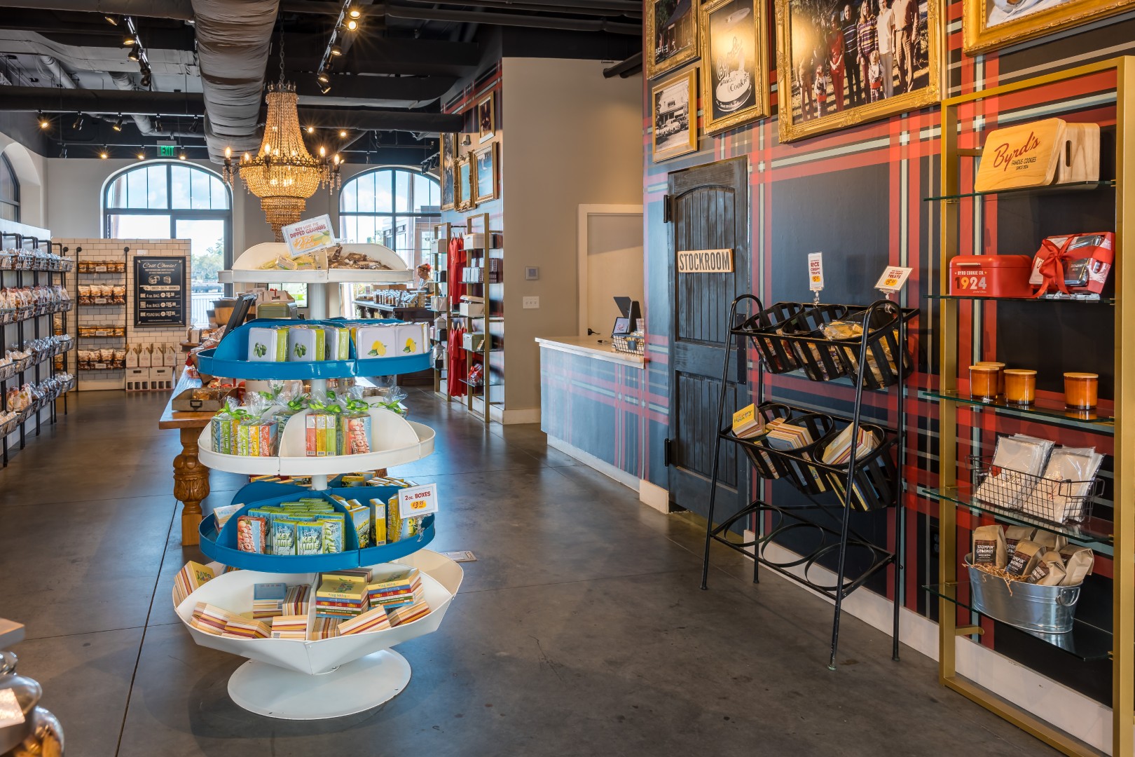 A well-lit gourmet shop interior with round shelves displaying packaged goods, racks with books and products on the right, and large windows and framed photos on the back wall.