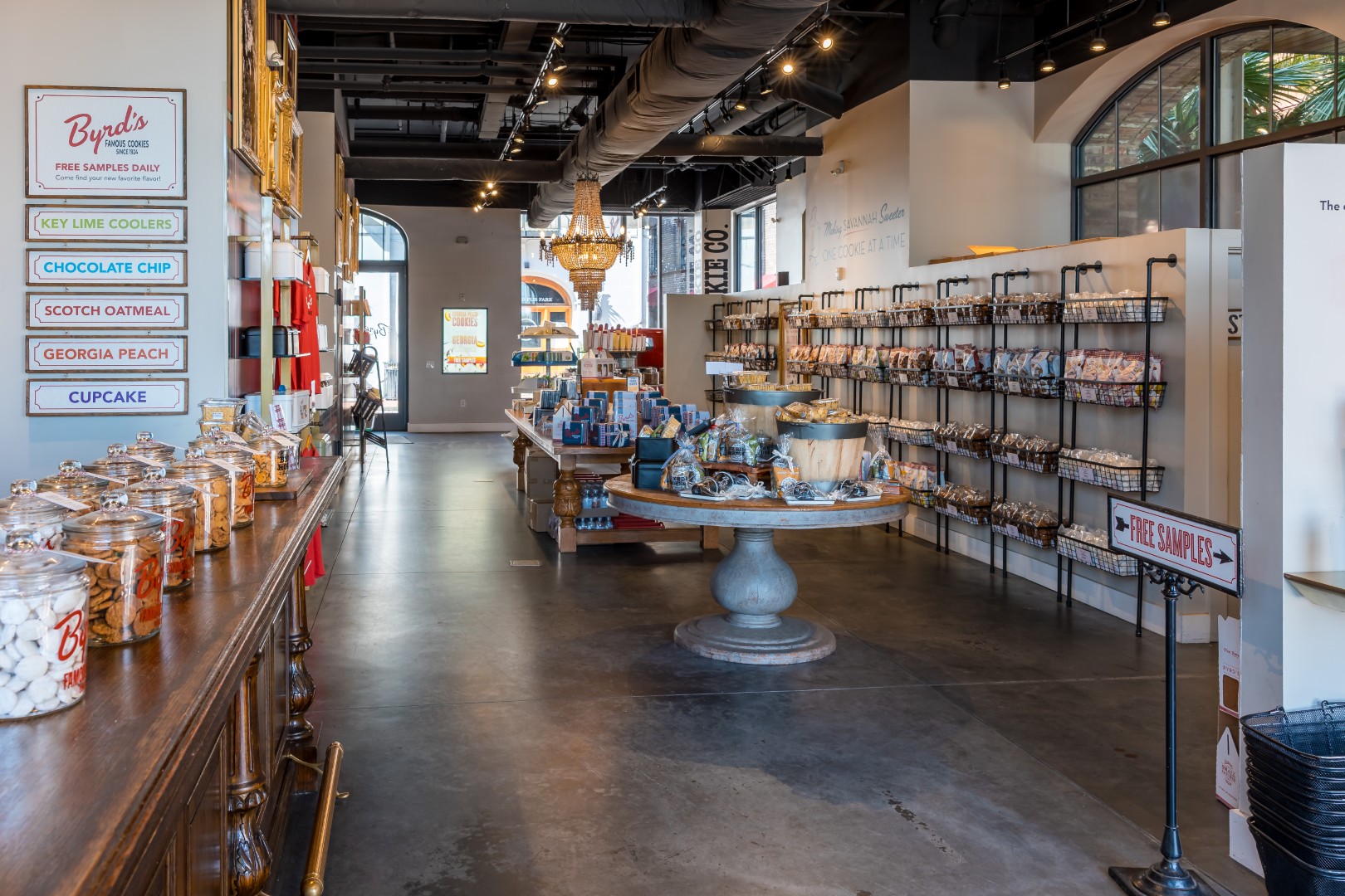 A bright, inviting cookie shop with glass jars of cookies on a wooden counter, shelves of packaged goods along the walls, and a central display table under a chandelier. Signs offer free samples and highlight various cookie flavors.