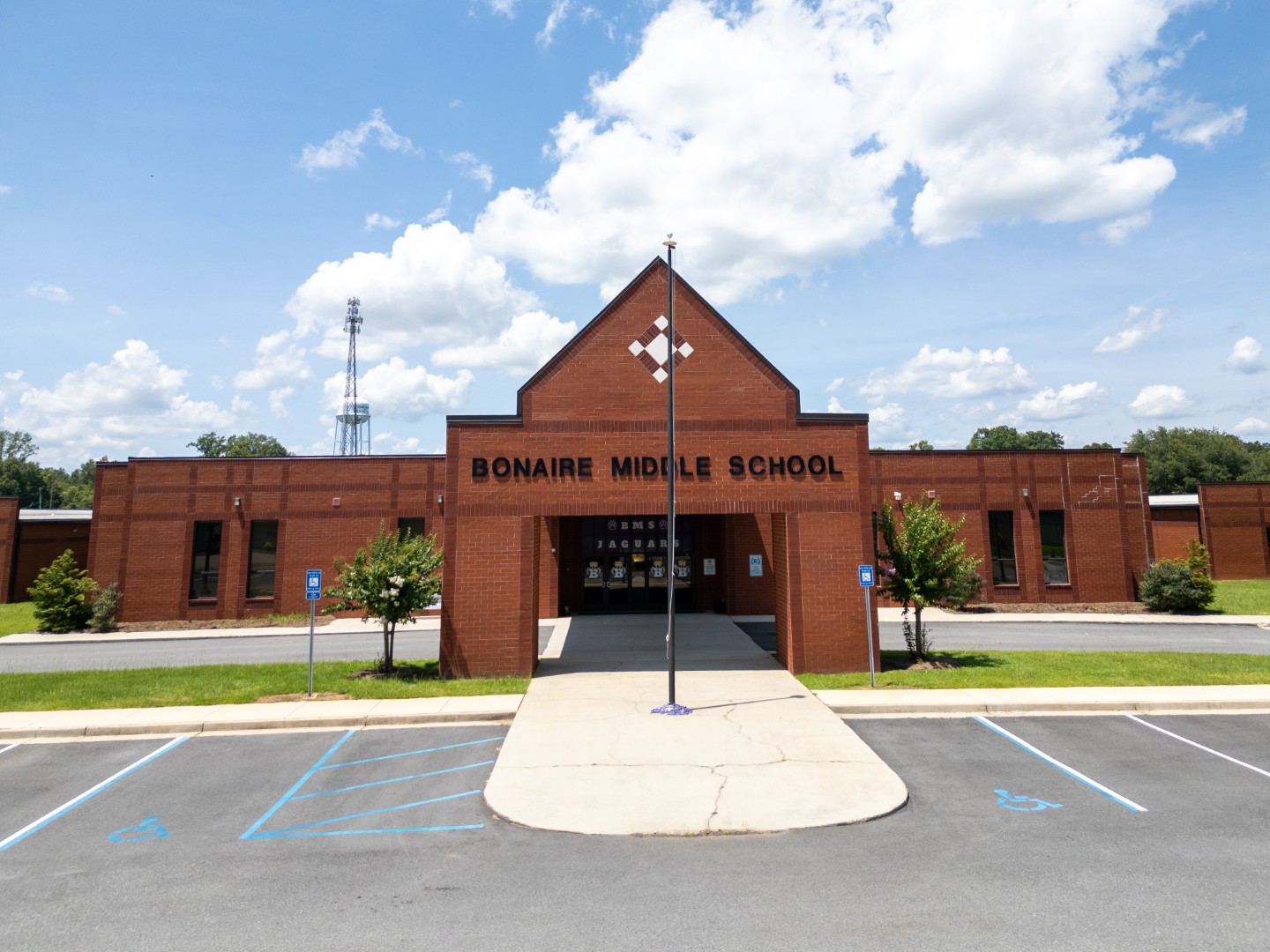 The front entrance of Bonaire Middle School, a red-brick building with a peaked roof, an American flag pole, small trees, and accessible parking spaces under a sunny sky with scattered clouds.
