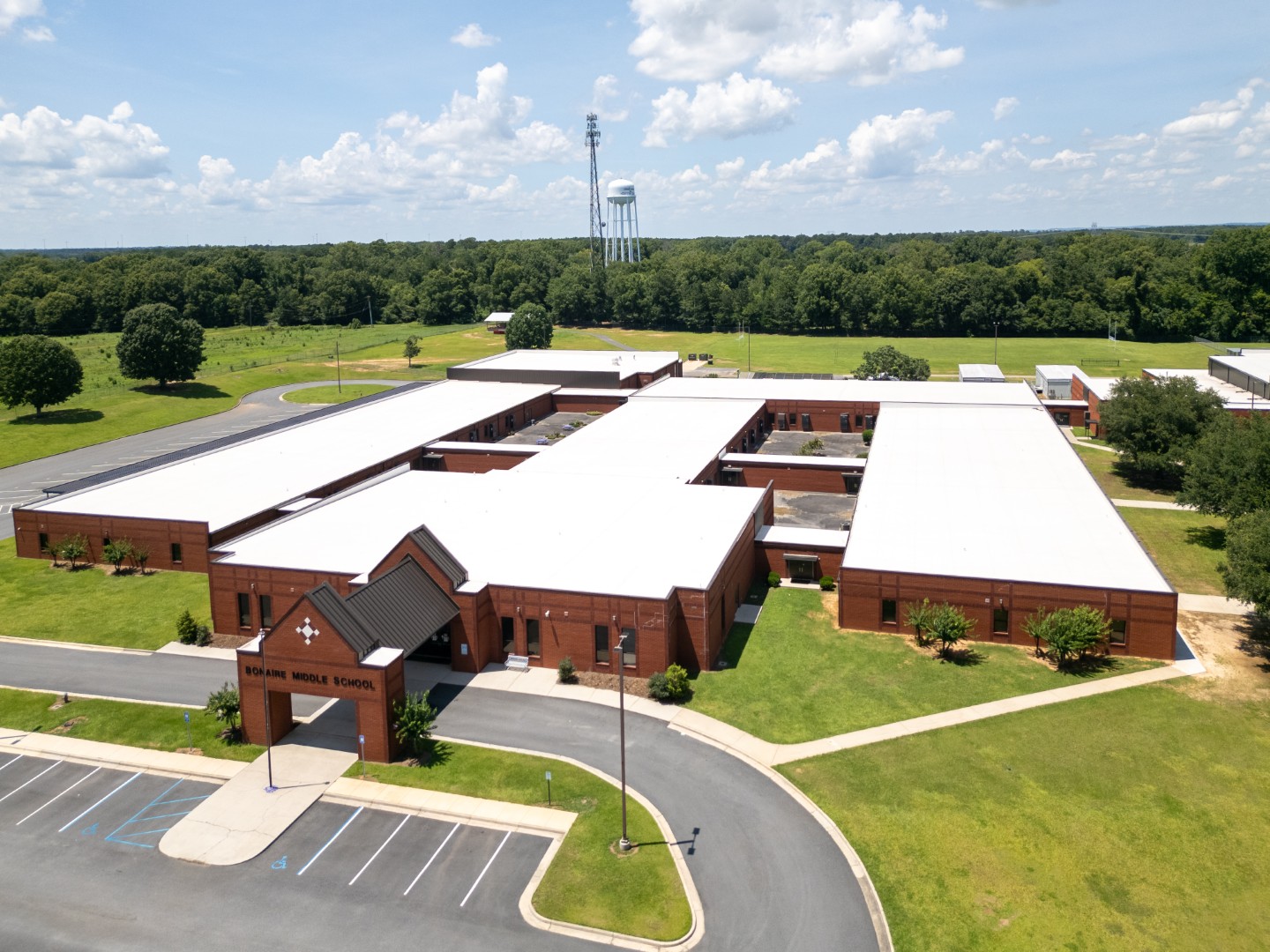 Aerial view of a large, single-story brick school building with white roofs, surrounded by green lawns, trees, a parking lot, and a distant water tower under a partly cloudy sky.