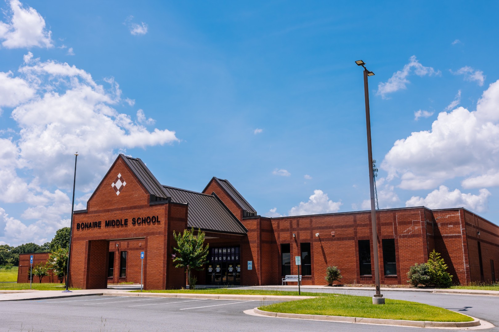 A red brick school building with a peaked roof labeled “Bonaire Middle School” under a blue sky with scattered clouds; the foreground shows an empty parking lot and some greenery.