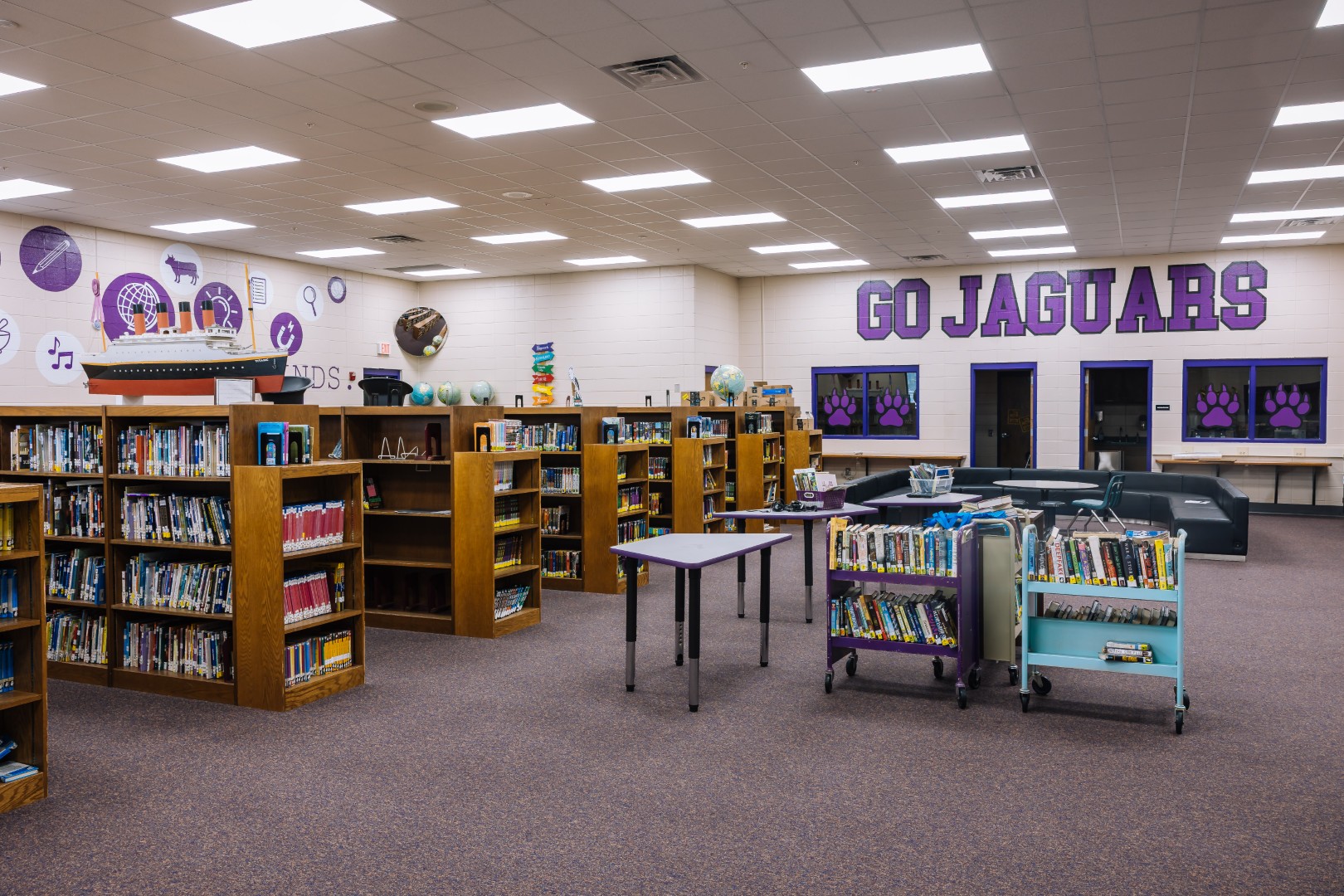 A school library with bookshelves, book carts, and tables. The back wall displays the words GO JAGUARS in large purple letters, with paw prints and educational murals on the walls.
