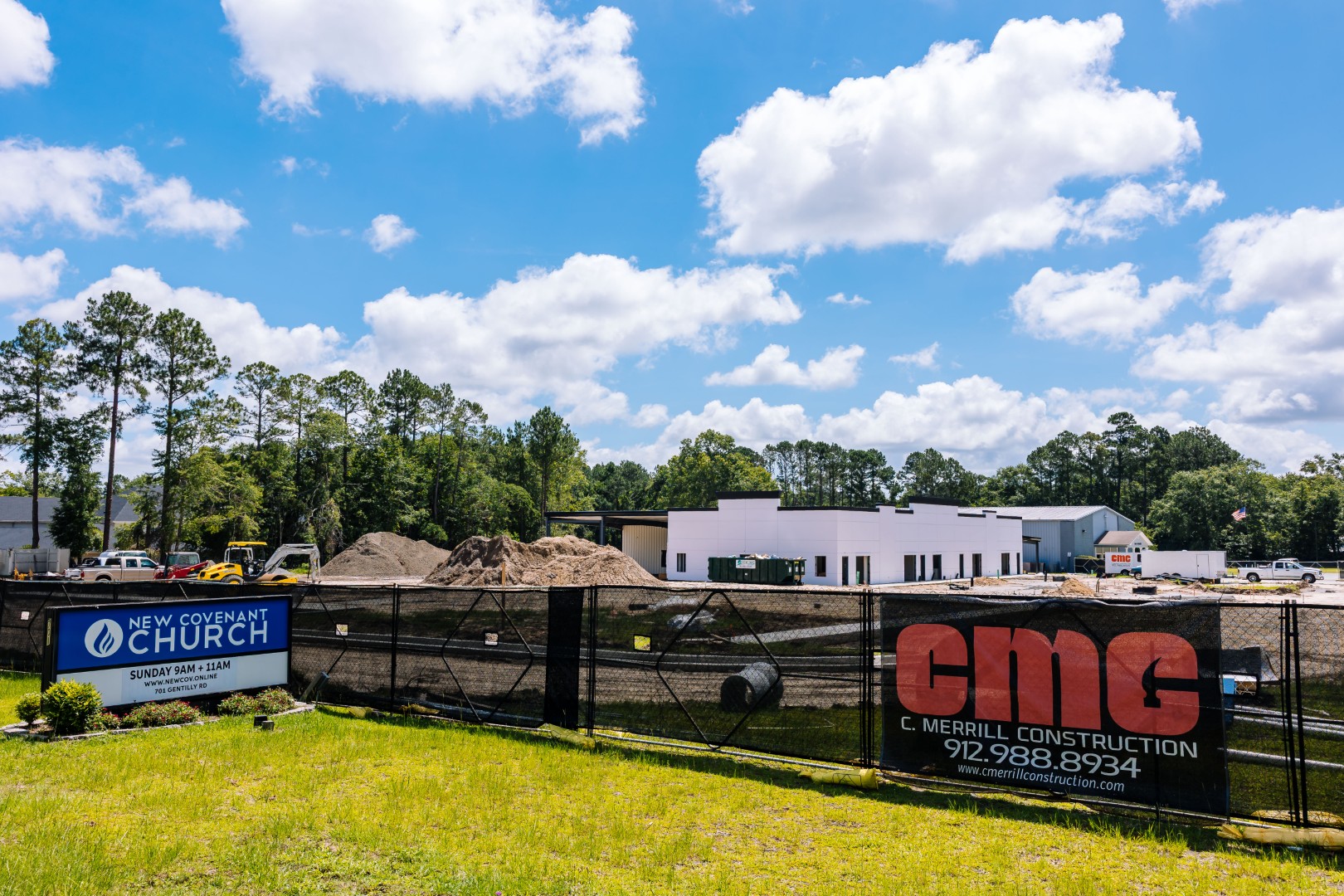 A construction site for New Covenant Church is visible behind a black fence with signs for the church and a construction company. Piles of dirt, construction vehicles, and a partially built building are in the background, under a blue sky with clouds.