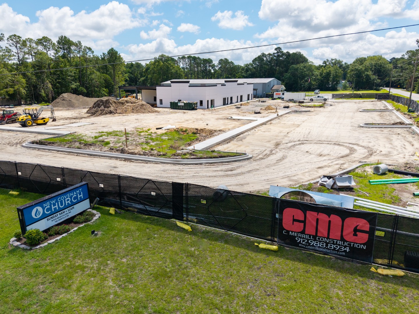A construction site with earth-moving equipment and a partially built structure in the background; signs on the fence read “New Covenant Church” and “C. Merrill Construction.” Trees and a blue sky are visible behind the site.