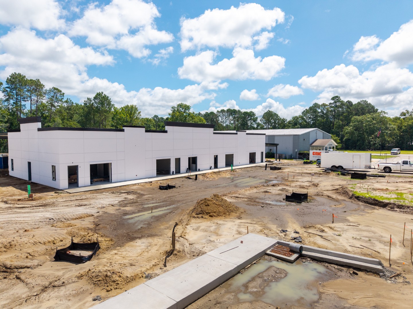 A partially constructed commercial building with white exterior walls and no doors or windows installed, surrounded by muddy ground, construction materials, and equipment under a partly cloudy sky.