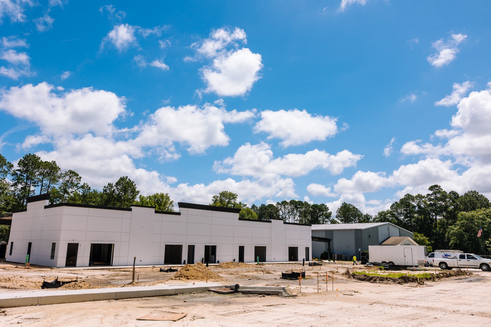 A white commercial building under construction, surrounded by dirt, construction materials, and a few vehicles, with trees and a bright blue sky with scattered clouds in the background.
