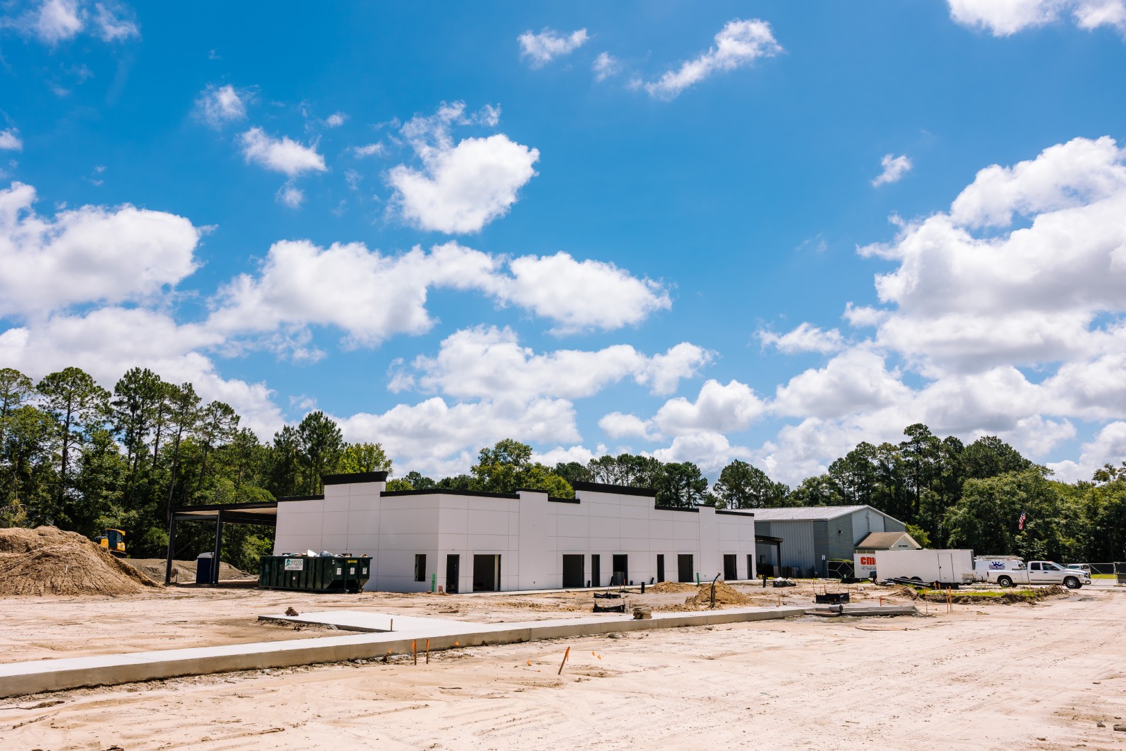A white commercial building under construction stands on a sandy lot under a bright blue sky with scattered clouds, surrounded by trees and construction materials.