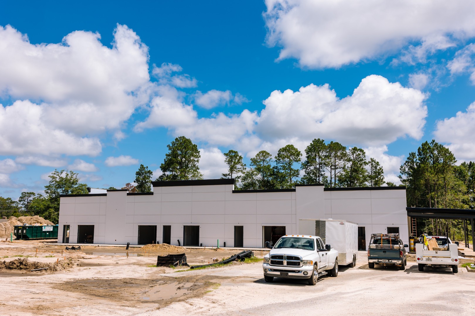 A modern white commercial building under construction, with work vehicles and trailers parked in front, surrounded by dirt, sand, and scattered construction materials. Tall trees and a bright blue sky with scattered clouds are in the background.