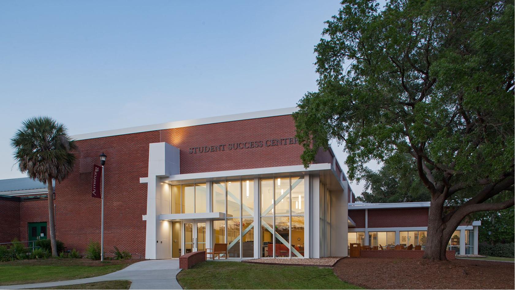 A brick building labeled Student Success Center with large glass windows, a tree and a palm tree nearby, and a curved sidewalk leading to the entrance at dusk.