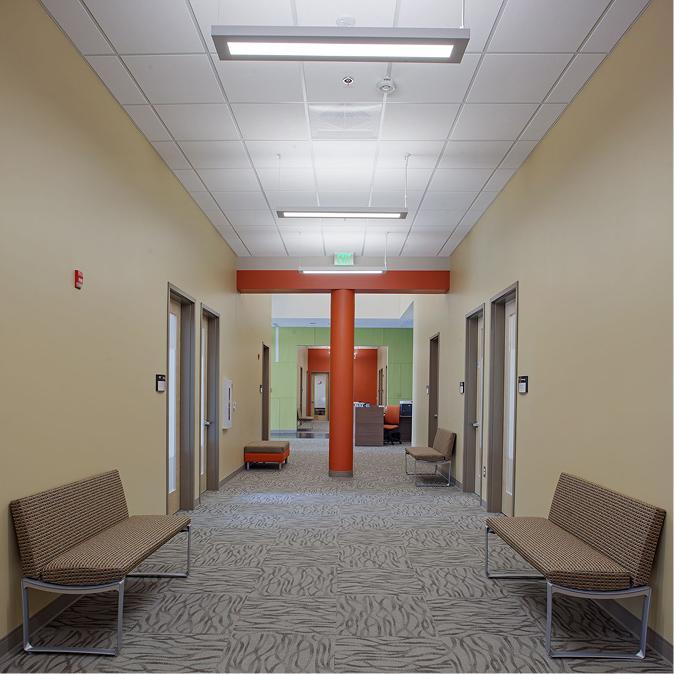 A hallway with beige walls, patterned carpet, benches along each side, and doors leading to offices. Bright overhead lighting and an orange accent wall are visible in the background.