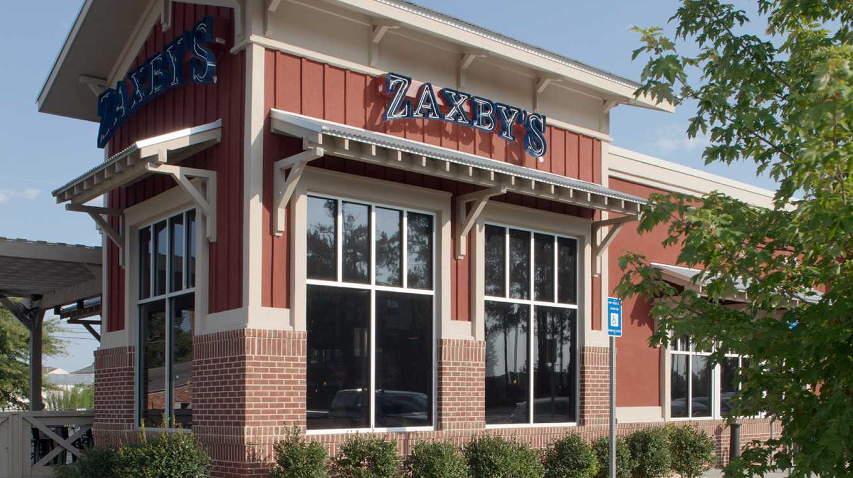 A Zaxby’s restaurant with red siding, large windows, and a brick foundation, partially shaded by trees, on a sunny day.