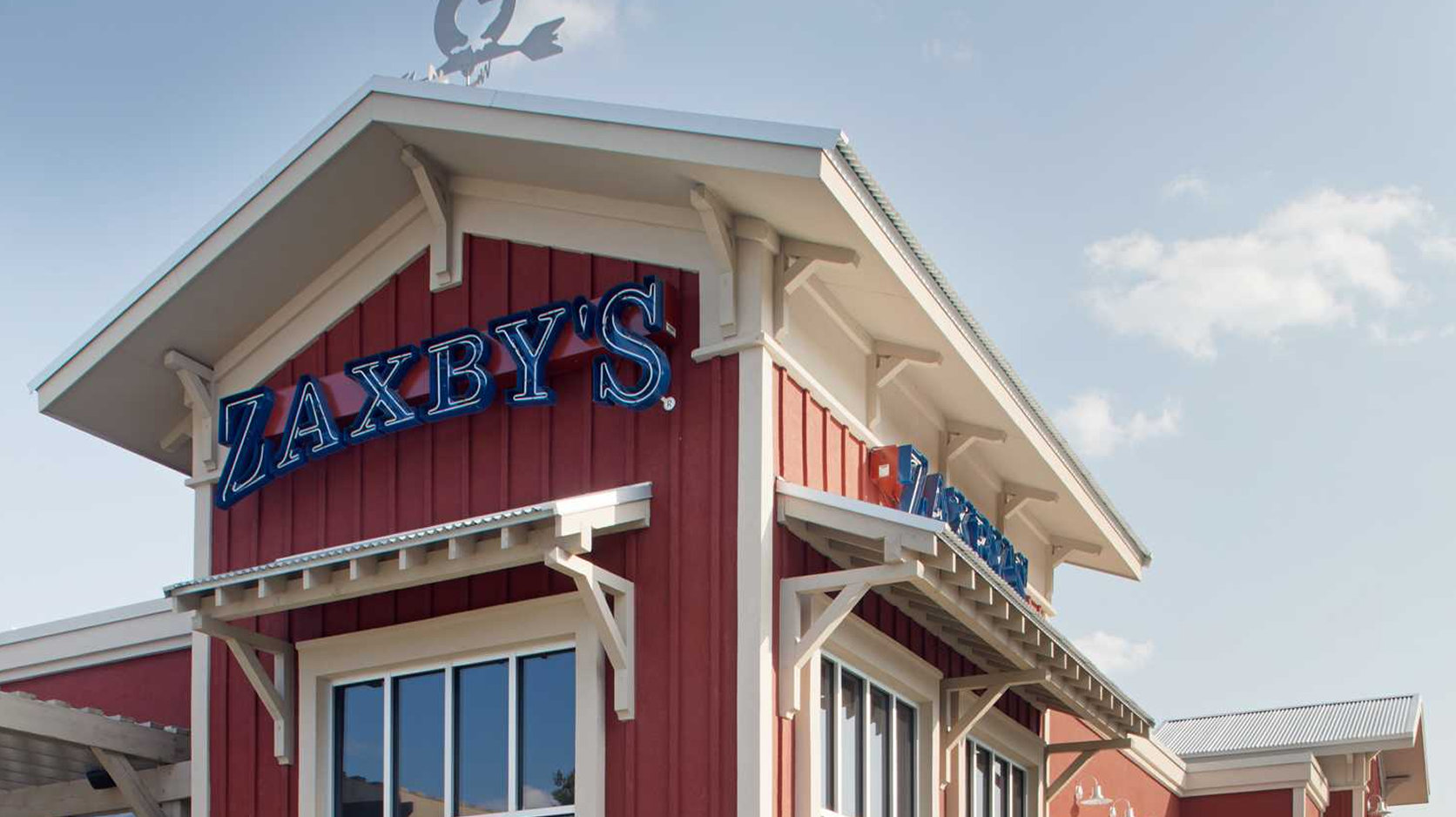 The image shows the exterior of a Zaxby's restaurant with a red and white facade, large windows, and a blue sky in the background. The Zaxby's sign is prominently displayed on the building.