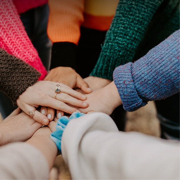 A group of people wearing colorful sweaters place their hands together in the center, showing unity and teamwork.