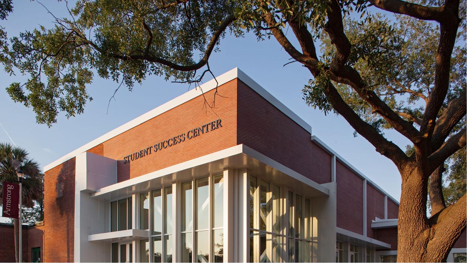 A brick building labeled Student Success Center with large glass windows, surrounded by leafy trees and bathed in warm sunlight.