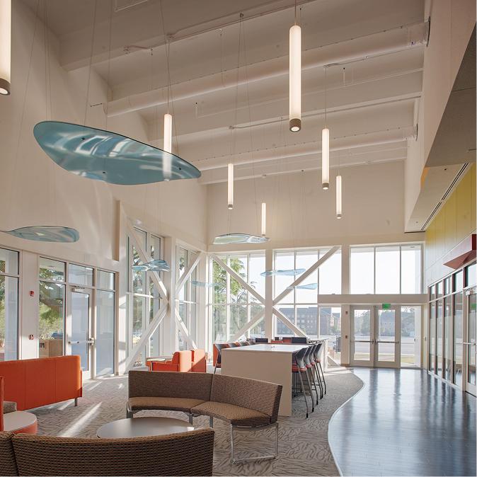 A sunlit lounge area with large windows, orange and beige seating, high tables with chairs, and hanging blue decorative panels on the ceiling. Vertical pendant lights add a contemporary feel.