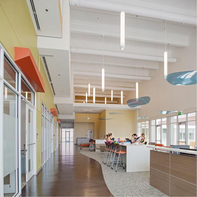 A brightly lit communal workspace with high ceilings, large windows, hanging lights, and people working at tall tables with orange chairs.