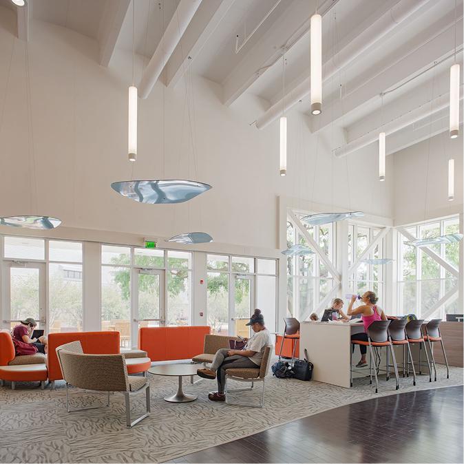 A bright lounge with large windows, orange and beige seating, bar stools at a counter, and people working or relaxing in natural light under hanging lights.