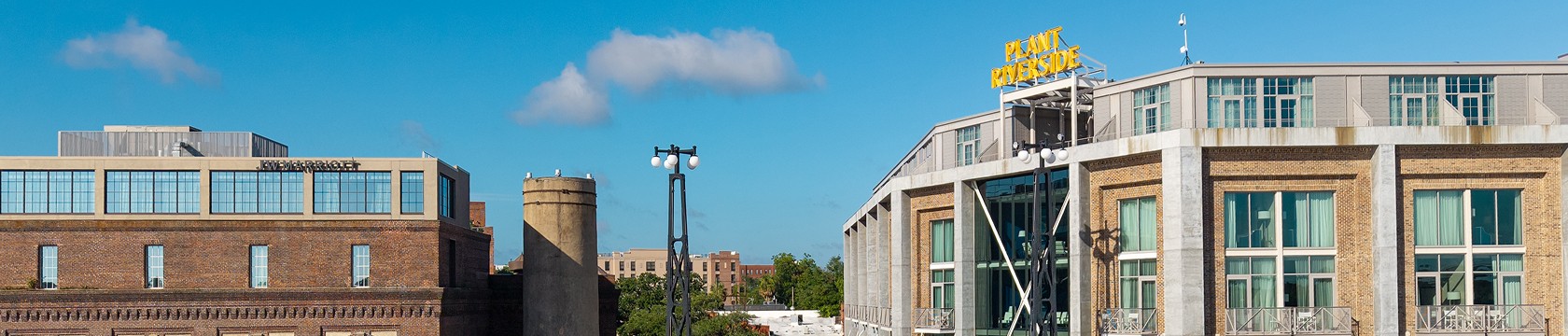 A panoramic view of industrial-style brick buildings with large windows under a blue sky; one building features a rooftop sign that reads PLANT RIVERSIDE. Trees and distant buildings are visible in the background.